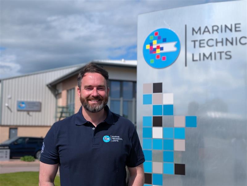 Smiling man stands in front of company logo in bright sunshine.