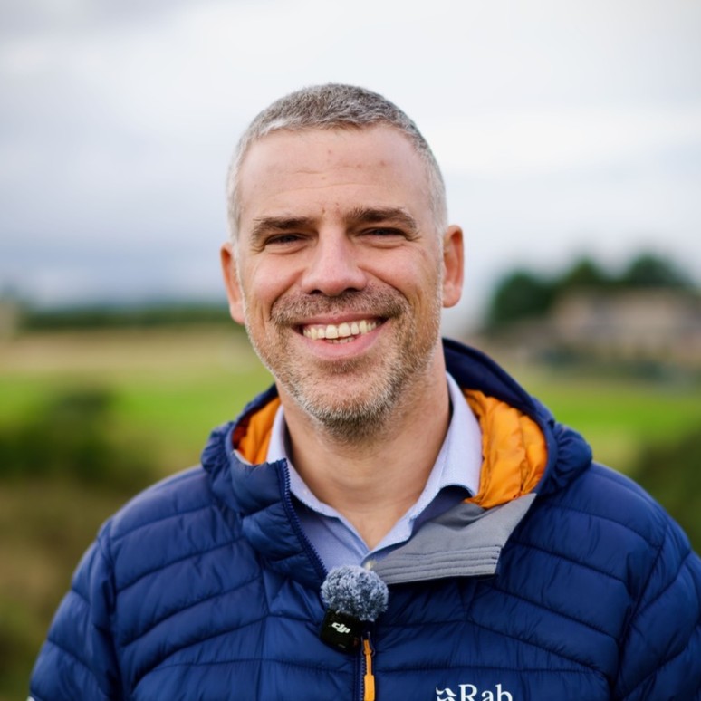 Portrait of Grigorios Gryllias outdoors, wearing a blue insulated jacket with a clip-on microphone, photographed against a softly blurred rural background.