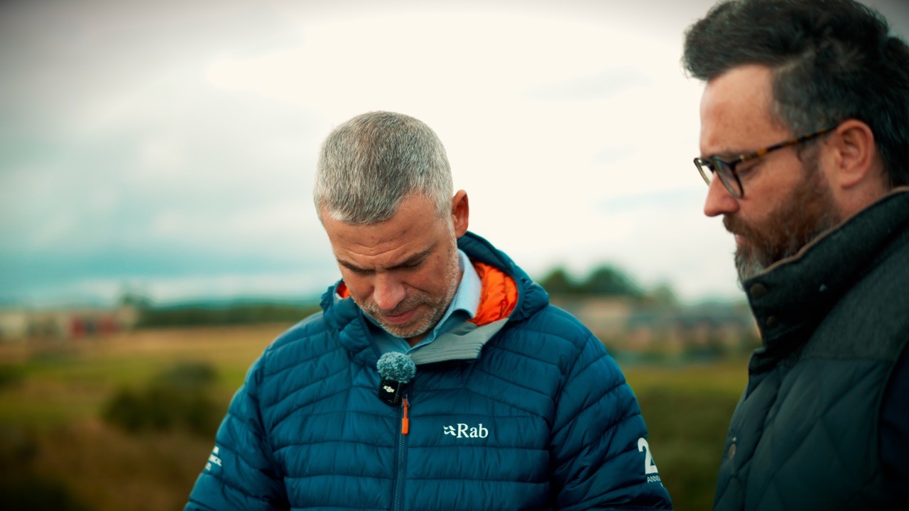 Two people standing outdoors during a filmed discussion, with one person wearing a blue insulated jacket and clip-on microphone. The background shows an open landscape under an overcast sky with a shallow depth of field