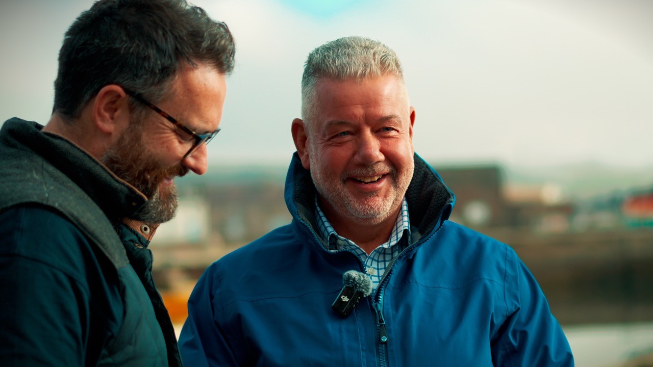 Two MTL colleagues during a filmed conversation, standing outdoors at a harbour location in Aberdeen with the boats softly out of focus in the background.