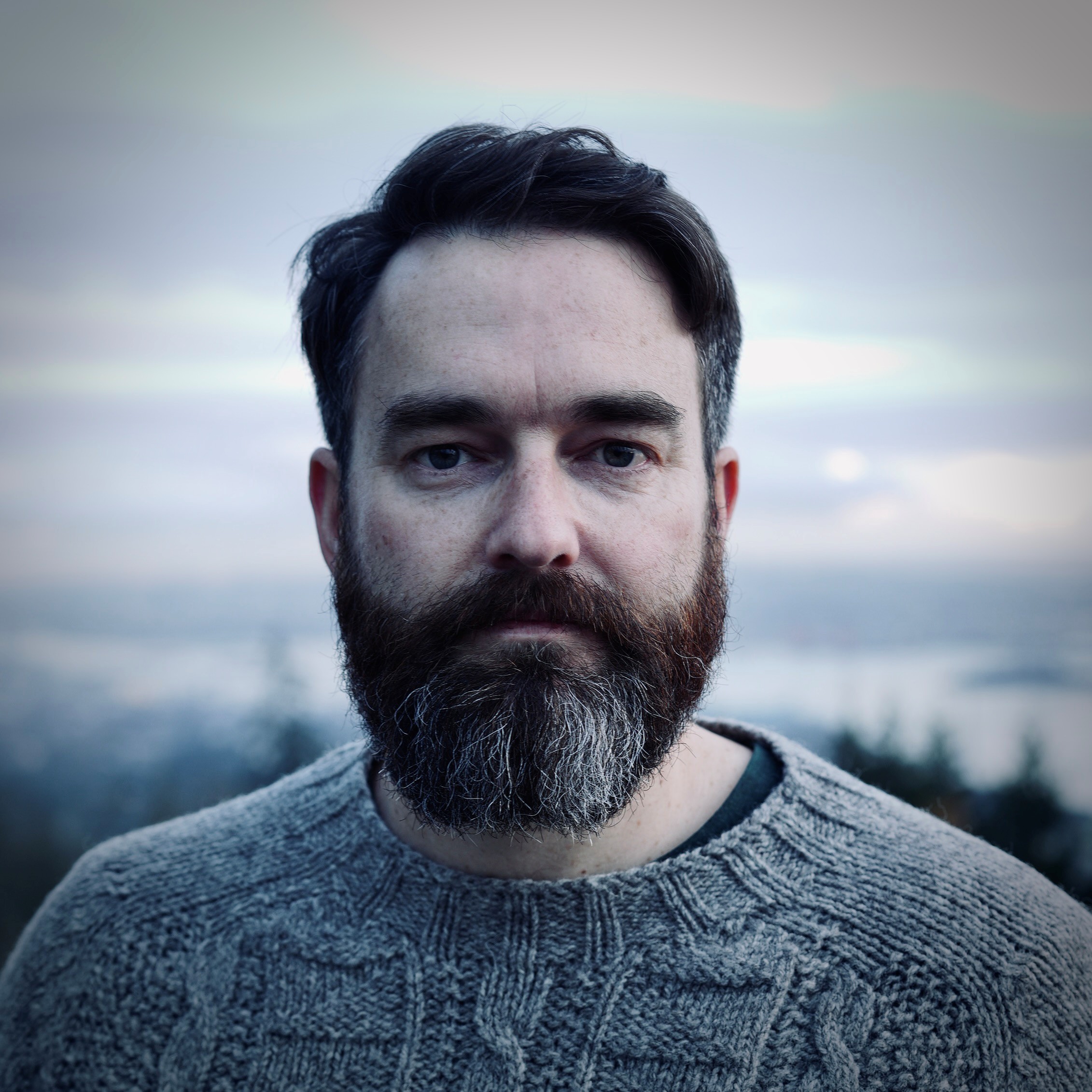 Close-up portrait of a bearded man wearing a grey knitted jumper, photographed outdoors with a blurred landscape and cloudy sky in the background